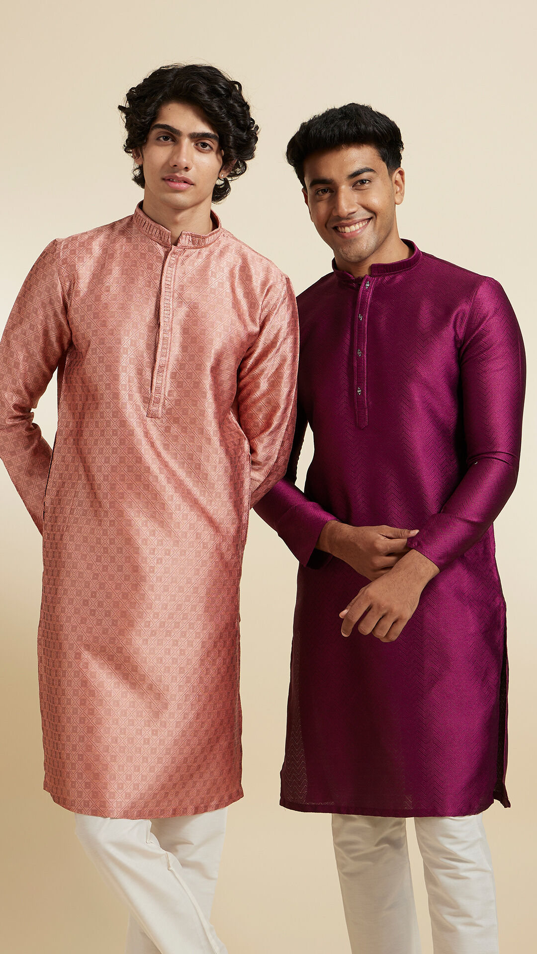 Two young men in traditional ethnic wear, one in a peach kurta and the other in a deep purple kurta, paired with white churidar pants, smiling and posing together.